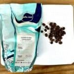 Roasted coffee beans next to a dotted notebook on a wooden surface, with a bag of Arabica Sundan Giling Basah coffee from Kafe Inc, highlighting premium coffee selection.