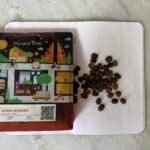 Golden roasted Arca Ulián washed coffee beans on a white textured surface, with colourful packaging featuring playful illustrations of a village scene and a QR code, promoting high-quality Bali coffee.