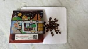 Golden roasted Arca Ulián washed coffee beans on a white textured surface, with colourful packaging featuring playful illustrations of a village scene and a QR code, promoting high-quality Bali coffee.