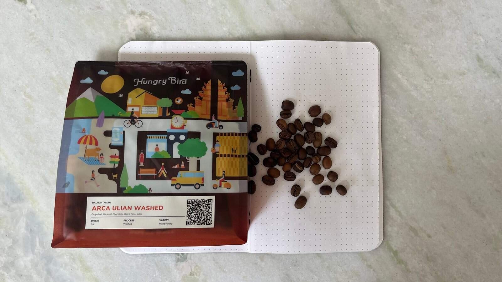 Golden roasted Arca Ulián washed coffee beans on a white textured surface, with colourful packaging featuring playful illustrations of a village scene and a QR code, promoting high-quality Bali coffee.