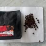 Rich coffee beans and a bag of Space Roastery’s Blood Orange chocolate nibs on a white textured surface.