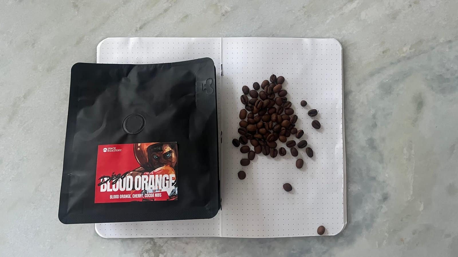 Rich coffee beans and a bag of Space Roastery’s Blood Orange chocolate nibs on a white textured surface.