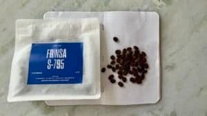 Roasted coffee beans next to a bag of Frinsa S-795 natural lactic ferments on a marble surface.