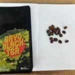 Rich black coffee beans on an open notebook with dotted pages, alongside a colourful "Gayo Black Tea" packet on a wooden surface, representing artisanal coffee and tea products.