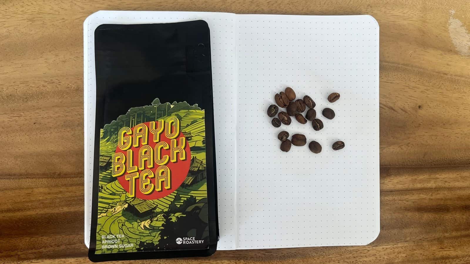 Rich black coffee beans on an open notebook with dotted pages, alongside a colourful "Gayo Black Tea" packet on a wooden surface, representing artisanal coffee and tea products.