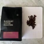 Rich coffee beans from Gueribe, Ethiopia, on a white perforated surface next to a bag of "Blacklist Coffee Roasters" fresh coffee. Perfect for brewing high-quality, speciality coffee at home.