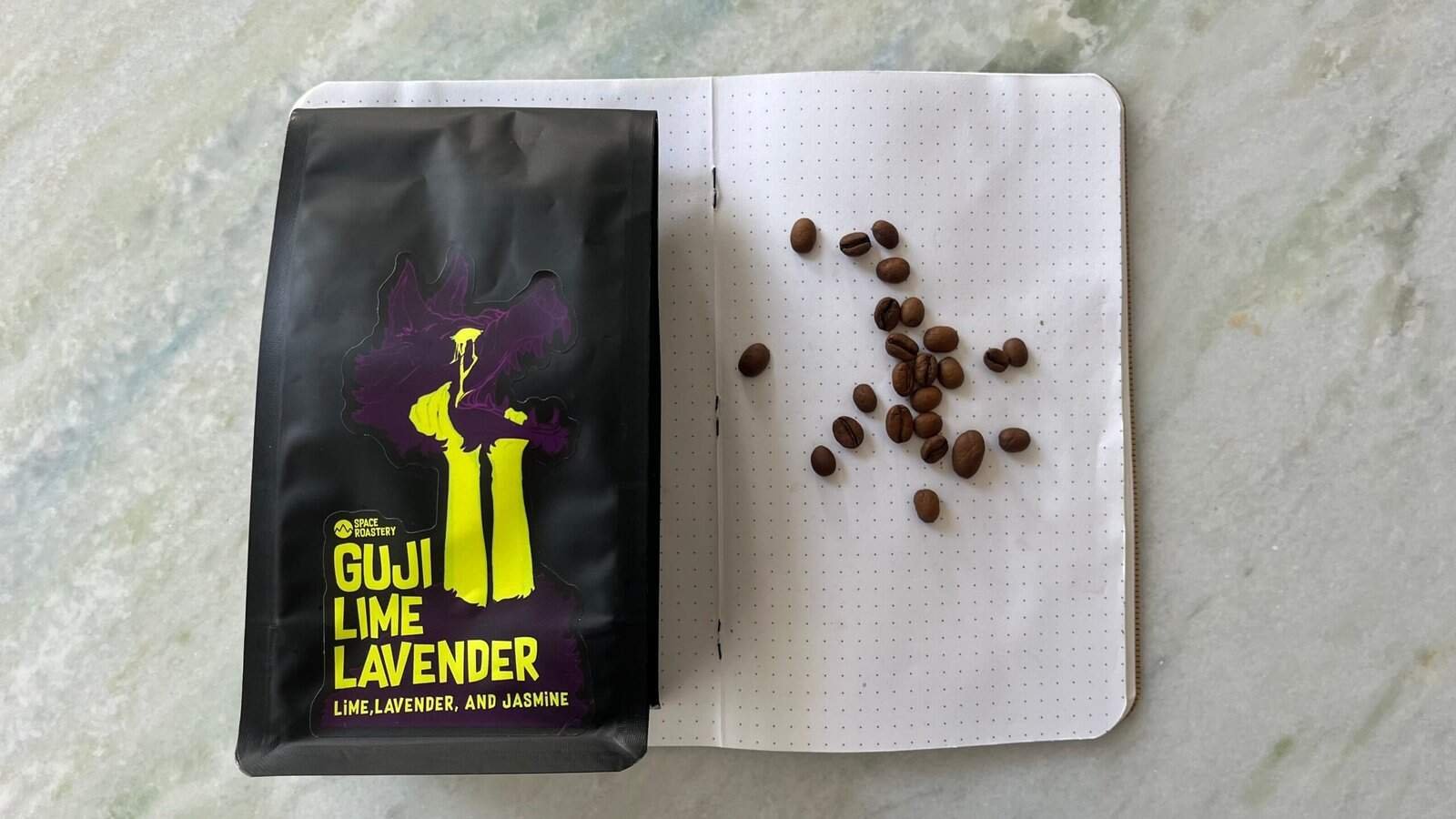 Rich dark roast coffee beans with Guji Lime Lavender branded packaging on a white dotted notebook. Coffee beans are spread out on the notebook, highlighting speciality coffee from Guji, Ethiopia.