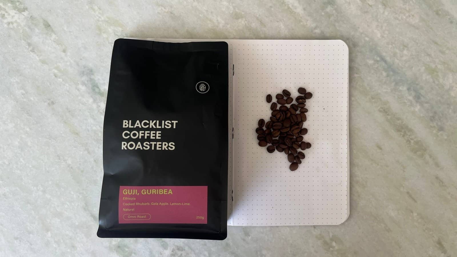 Rich coffee beans from Gueribe, Ethiopia, on a white perforated surface next to a bag of "Blacklist Coffee Roasters" fresh coffee. Perfect for brewing high-quality, speciality coffee at home.