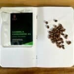 Roasted coffee beans on a dotted notebook page next to a package of Hibela Cardamom coffee, showing coffee beans and spices for artisanal coffee brewing.