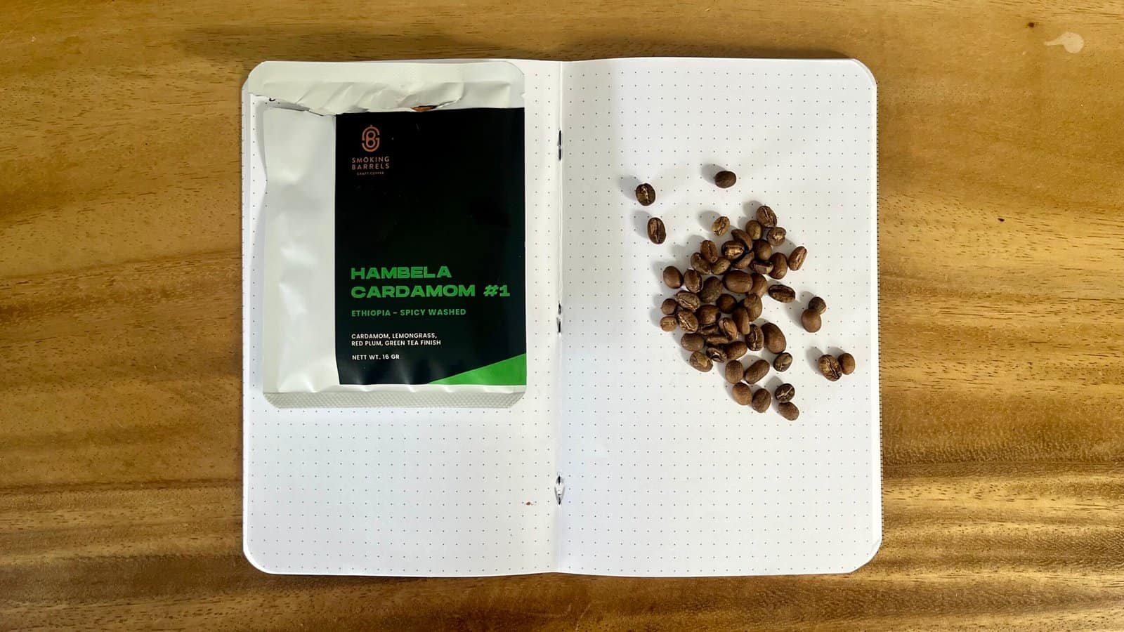 Roasted coffee beans on a dotted notebook page next to a package of Hibela Cardamom coffee, showing coffee beans and spices for artisanal coffee brewing.