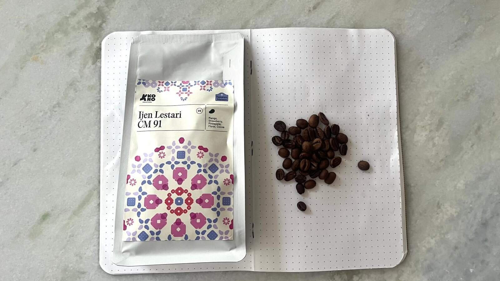 Rich coffee beans and a package of Ijen Lestari CM 91 specialty coffee, on an open dotted notebook, highlighting premium single-origin coffee for connoisseurs.