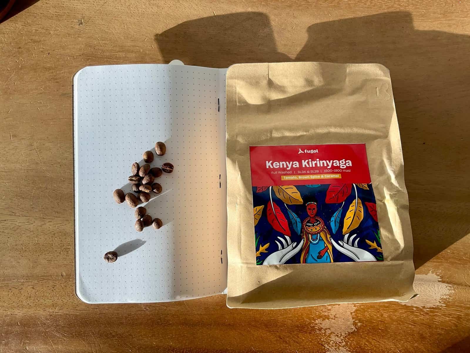 Realistic photo of Kenyan coffee beans on a notebook page with a bag of Kenyan Kirinyaga coffee on a wooden surface, highlighting premium coffee sourcing and quality beans.