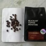 Rich, dark coffee beans spilled on a notebook next to a bag of ブラックリストコーヒーロースターズ Papayo Bourbon coffee, highlighting speciality beans for espresso lovers.