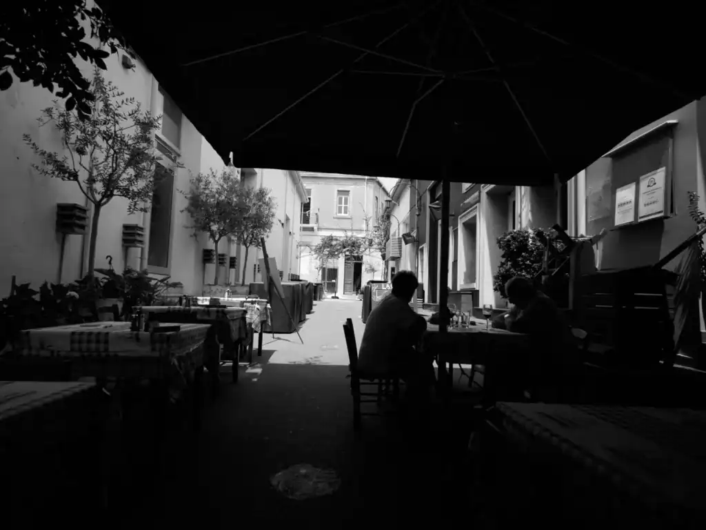 A black and white photo of an outdoor restaurant patio with tables and chairs under a large umbrella, trees lining the narrow alleyway, and two people sitting at a table, creating a cosy atmosphere.
