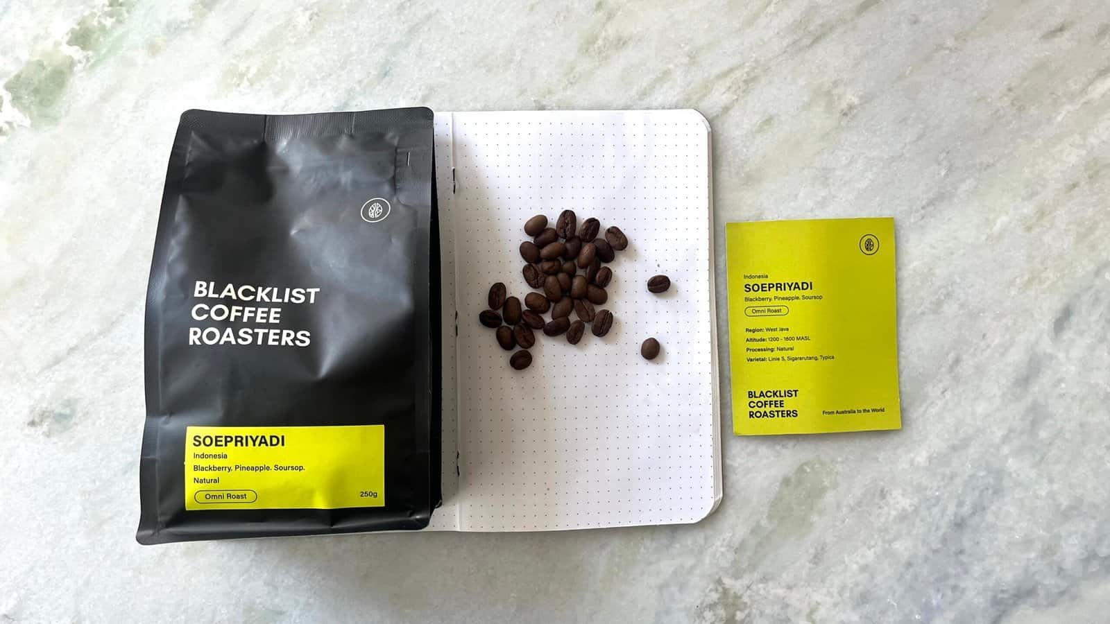 Bright yellow and black coffee packaging with roasted coffee beans on a white dotted notebook, showcasing premium Indonesian Blacklist coffee beans, highlighting speciality coffee beans and sustainable sourcing.