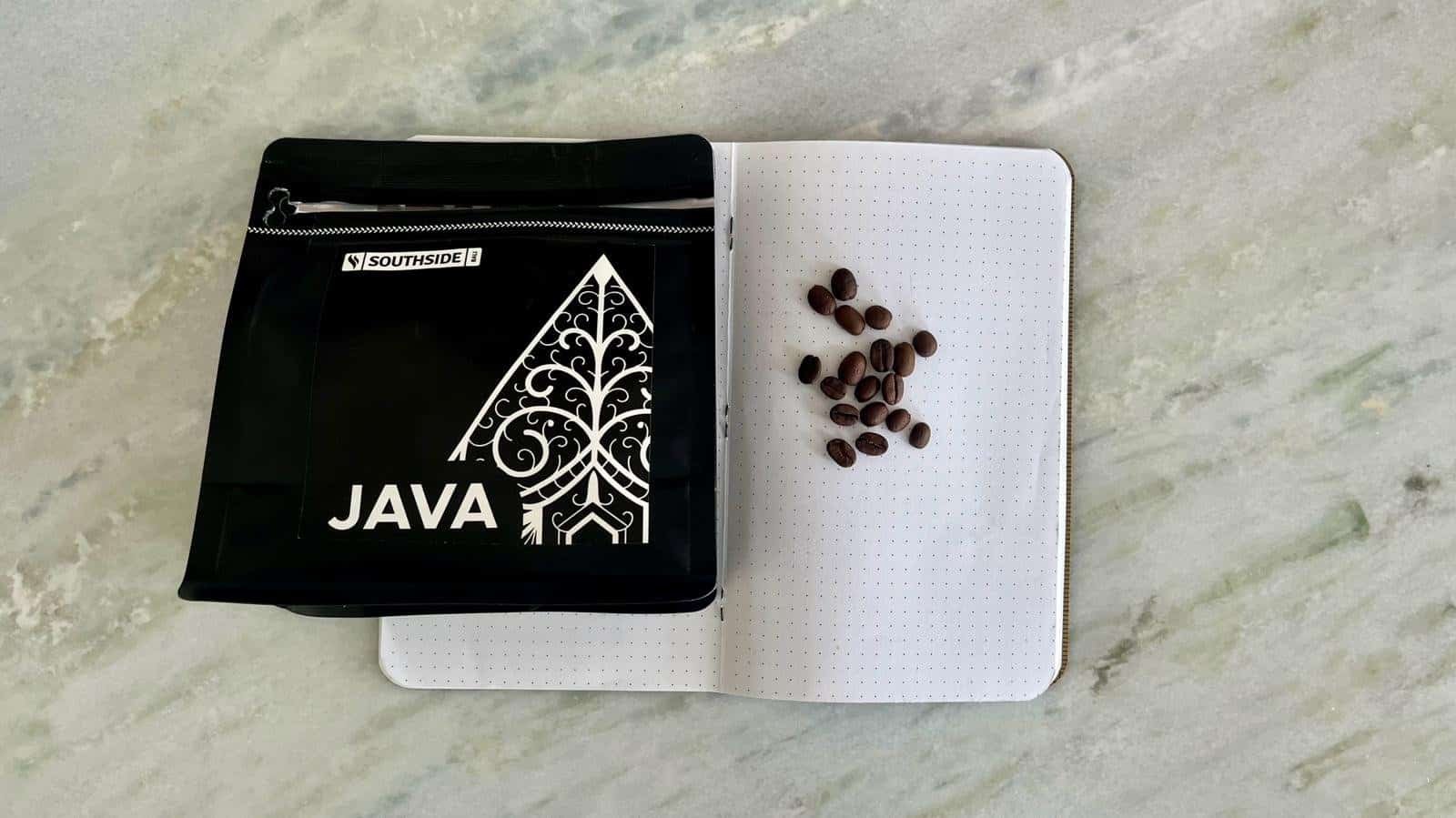 Coffee beans scattered on dotted notebook next to a black Java programming book with an intricate design.