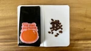 Rich coffee beans and a bag of "The Pink Mandarín" speciality coffee on a notebook with coffee beans arranged in a pattern, showcasing premium coffee roasting by Barnaby Robson.