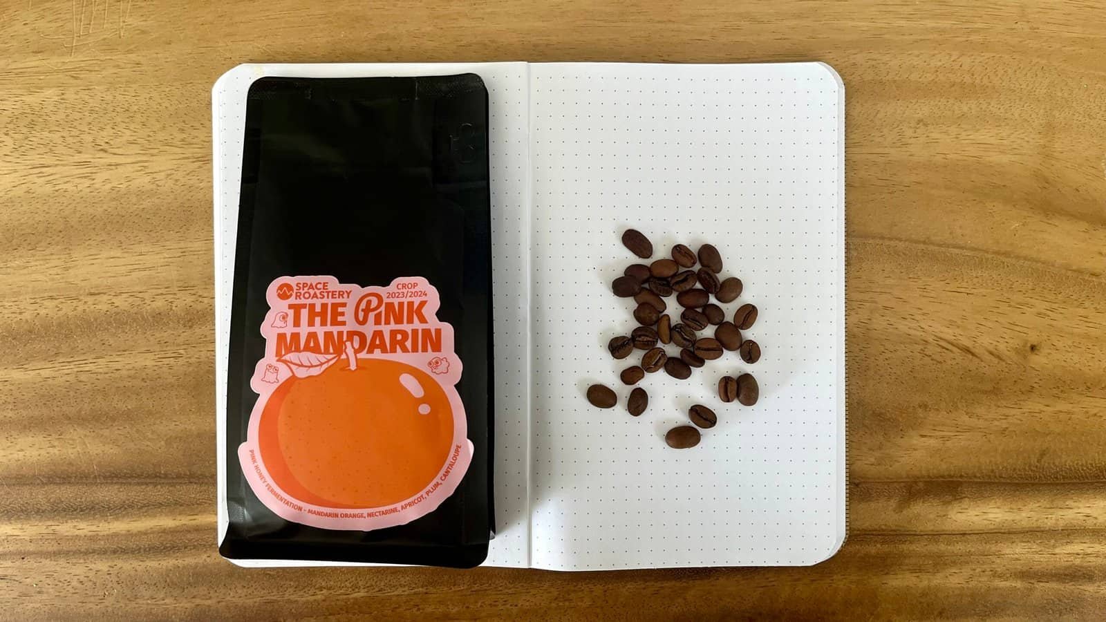 Rich coffee beans and a bag of "The Pink Mandarín" speciality coffee on a notebook with coffee beans arranged in a pattern, showcasing premium coffee roasting by Barnaby Robson.