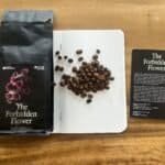 Rich roasted coffee beans from Barnaby Robson’s The Forbidden Flower blend on a dotted notebook, beside the product packaging and an informational card, highlighting artisanal coffee, single origin, and flavour profiles.