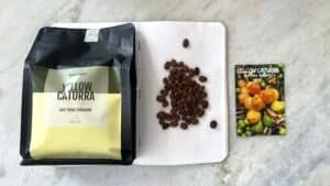 Dark roasted Yellow Caturra coffee beans from East Nusa Tenggara, Indonesia, displayed on a notebook with a colourful coffee farm card for organic coffee lovers.