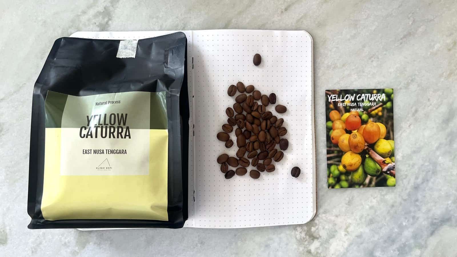 Dark roasted Yellow Caturra coffee beans from East Nusa Tenggara, Indonesia, displayed on a notebook with a colourful coffee farm card for organic coffee lovers.
